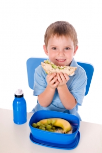 Boys WFL Smiling boy eating sandwich with a blue plastic lunchbox containing a banana and some grapes and a blue plastic water bottle