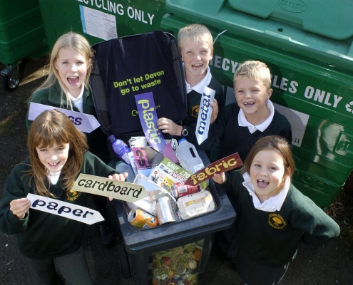 5 children around a bin filled with household rubbish holding signs that have different materials written on them.