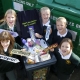 5 children around a bin filled with household rubbish holding signs that have different materials written on them.