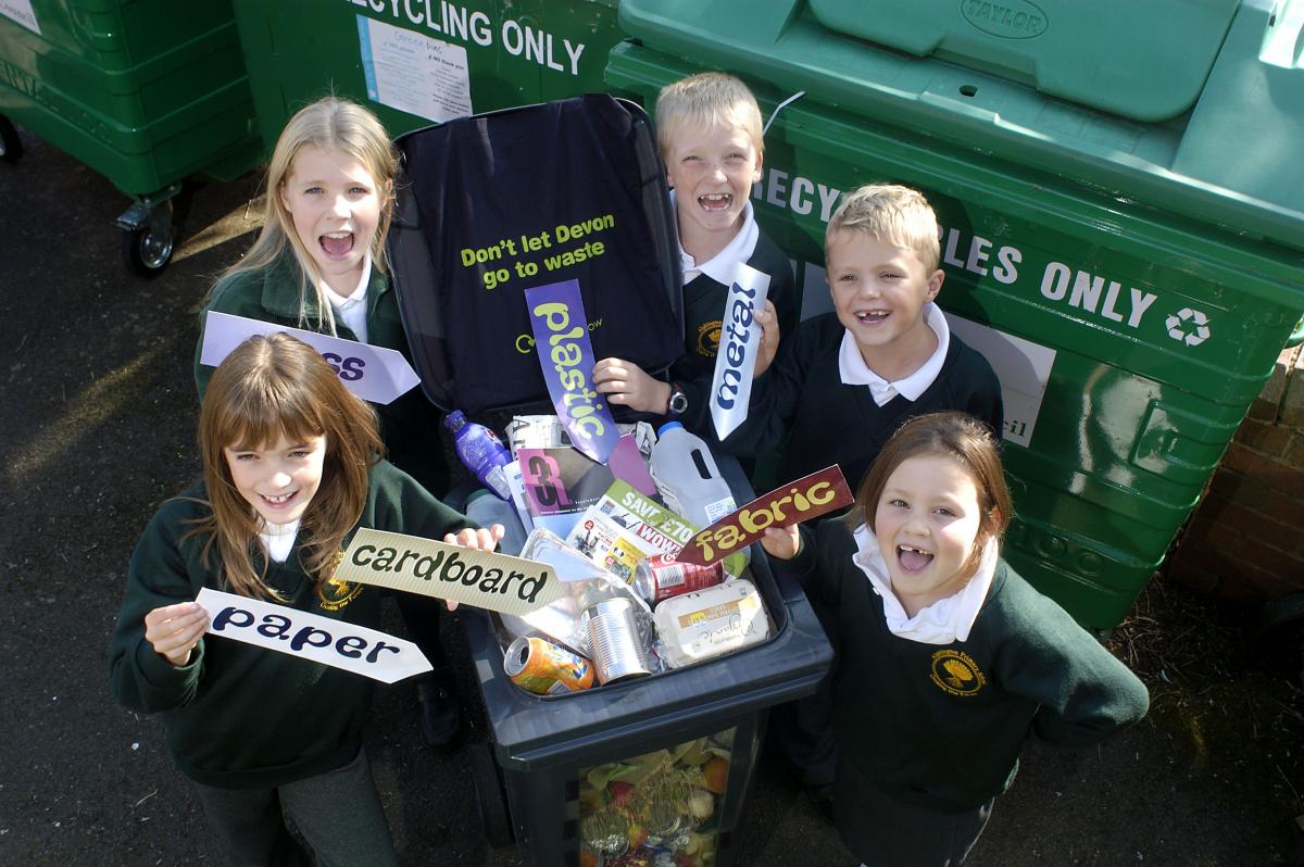 Children with recyclable material labels 5 children around a bin filled with household rubbish holding signs that have different materials written on them.