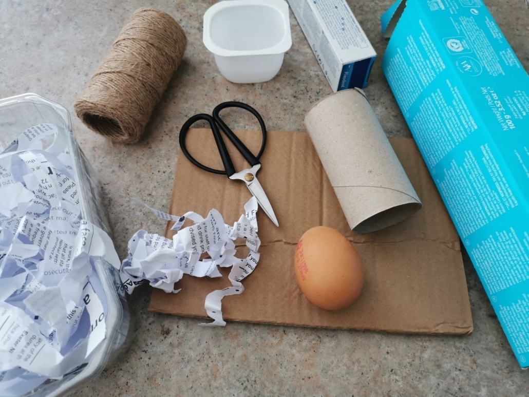 Egg drop activity An egg is surrounded by empty cardboard packaging, a yogurt pot, a fruit punnet, shredded paper, scissors and string.