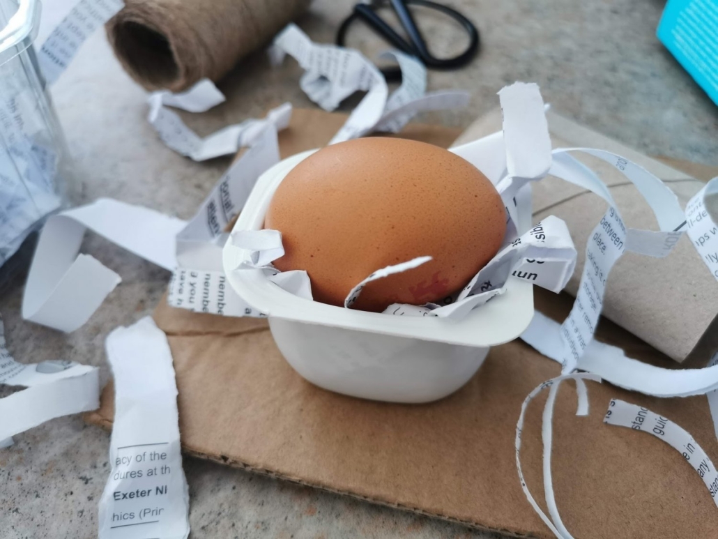 Egg drop activity 2 An egg rests in a yogurt pot filled with shredded paper. There are various recyclable materials in the background of the image, including cardboard along with scissors and string.