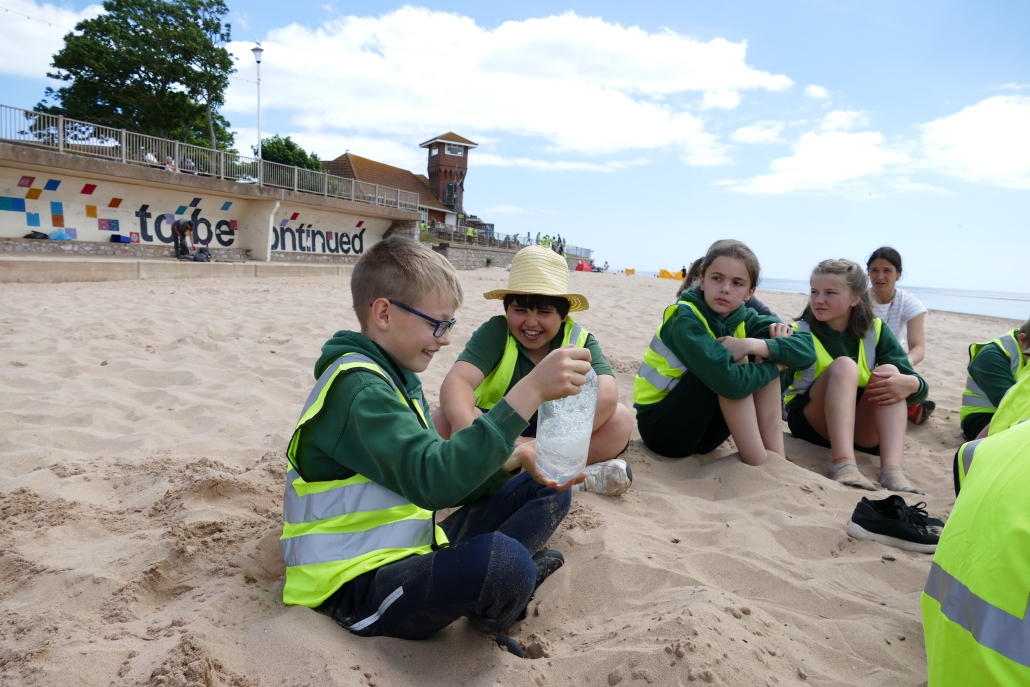 Four children sat on the sand, one holding a clear plastic bottle of water with plastic inside