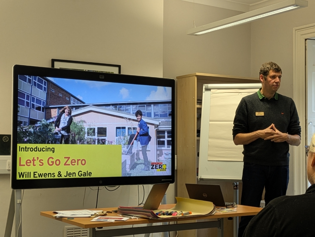 A man presenting in front of a TV showing PowerPoint slides about Let's Go Zero