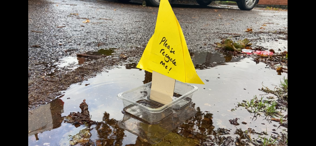 Plastic boat A transparent plastic pot with a piece of cardboard and papaer. The yellow paper saying 'please recycle me'. It is an example submission to the Big Recycling Hunt.