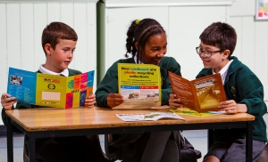 Poetry 3 happy children in green school uniform sat at a table reading leaflets about waste and recycling