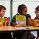3 happy children in green school uniform sat at a table reading leaflets about waste and recycling