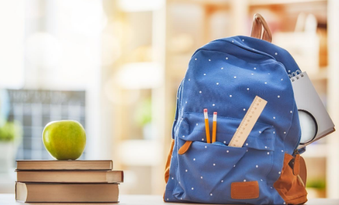 A stack of three books with a green apple on top, next to a blue spotted school rucksack, with two pencils and a ruler sticking out from the pocket.