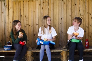 PSHE & Life Skills 3 smiling children sitting apart in an outdoor covered seat seat eating packed lunches