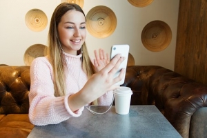 girl-on video call - pixabay A young woman wearing earphones is sitting at a table with a coffee cup. She holds her phone up to her face and smiles at the screen and waves.
