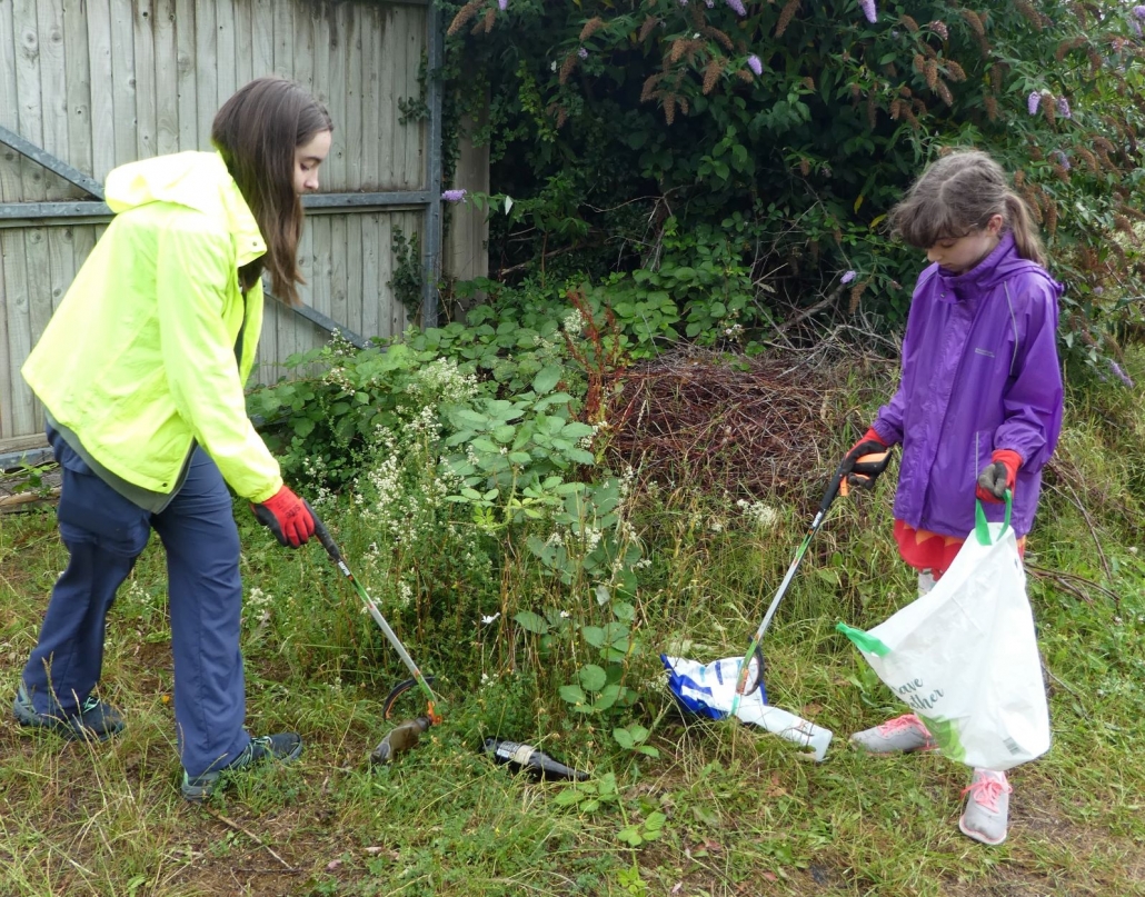litter picking 2 Girls wearing gloves picking up bottles and crisp packets with litter pickers from a bush.