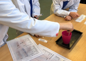 wetness test A photo of children in lab coats taking water from a cup using a pipette. This is with a recording sheet. You can't see the children's faces as its focused on their hands.